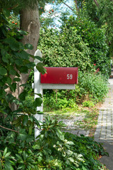 Red letterbox mounted on a white post surrounded by green bushes and trees