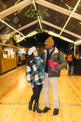 Young multi ethnic couple holding christmas gifts at a christmas market illuminated with christmas...