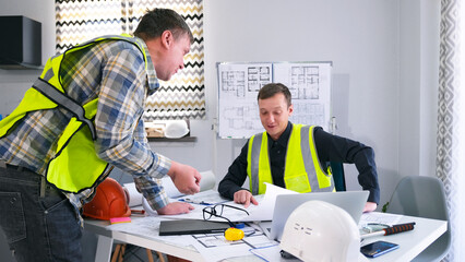 Two workers in green signal vests are discussing construction plans. The guys look at the drawings and discuss the details of the project