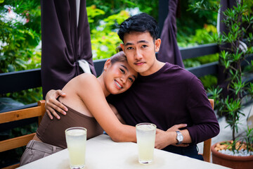 Smiling young Asian couple embrace while looking at the camera during a date at an alfresco cafe.