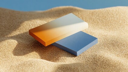 Two rectangular blocks, ombre colors (orange/yellow and blue), rest on textured sand.  Sunlight casts shadows