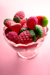 Glass bowl overflowing with sugary strawberry shaped gummy candy on pink background