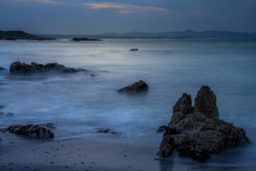 Rocky shoreline of Walker Bay at dawn, Hermanus, Western Cape, South Africa. Tranquil seascape with moody blue tones, misty water, and distant mountains under soft early morning light.