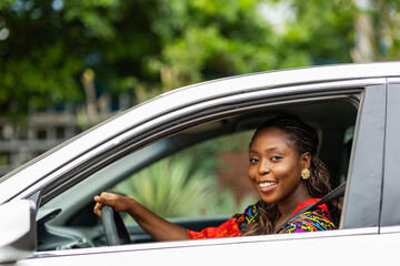 Smiling young Black African woman driving a white car in a neighborhood, looking at the camera.