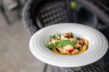 Healthy couscous salad with spinach, cherry tomatoes, grilled chicken, and roasted squash on a white plate outdoors on a wicker chair, close-up view.