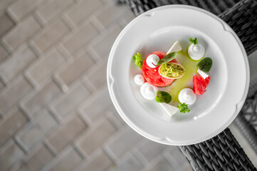 Gourmet summer salad with watermelon, avocado, cheese, and herbs, elegantly plated on a white dish against a textured backdrop.
