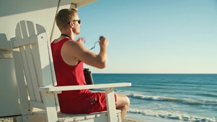 A male lifeguard in a red uniform sits in a white chair, watching the ocean from his tower on a sunny day.