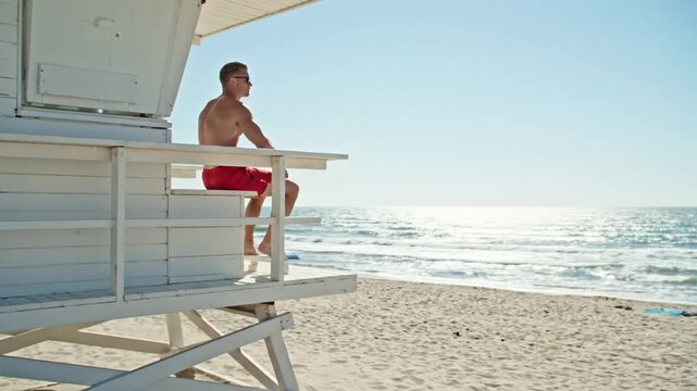 A shirtless lifeguard in red shorts sits on a white tower, watching over a sunny beach and sparkling ocean.