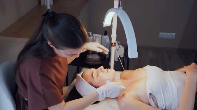 A cosmetologist in a beauty salon performs a skin cleansing procedure on a patient. The cosmetologist applies a mask to the woman's face to eliminate pigmentation and skin defects after acne.