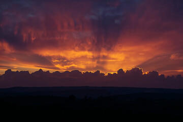 Fototapeta premium Fiery Sunset with Sun Rays Breaking Through Clouds Over Silhouetted Landscape dramatic