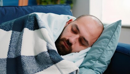 A close-up shot of a tired, bearded man sleeping deeply on a comfortable sofa, wrapped in a warm checkered blanket for rest.