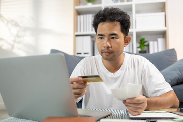 A man is sitting on a couch and looking at a receipt on a laptop. He is holding a credit card and he is checking his bank account. Concept of financial responsibility
