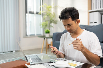 A man is sitting on a couch and looking at a receipt on a laptop. He is holding a credit card and...