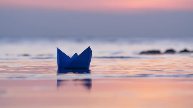 Blue paper boat stranded near shore during serene sunset hour, seen through telephoto lens. Ship rests motionless in shallow water, untouched by faint laps of calm sea waves.