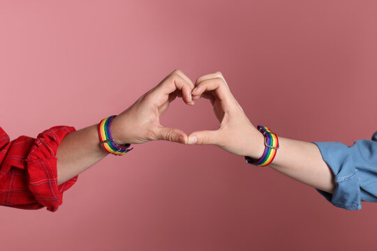 LGBT concept. Women in rainbow wristbands making heart shape on pink background, closeup