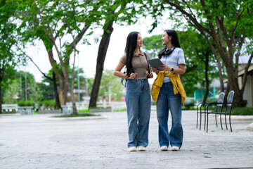 Two asian students walking and talking in a university campus park, holding a tablet