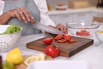 Senior woman cutting tomato at white marble table in kitchen, closeup
