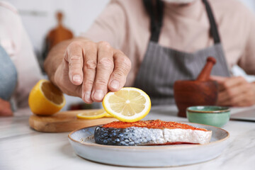 Elderly man putting slice of lemon onto salmon fish at white marble table indoors, closeup