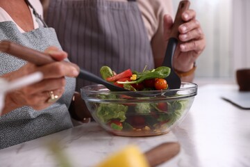 Elderly couple with spatulas cooking together at white marble table in kitchen, closeup