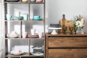 Shelving unit with different dishware, cups and floral decor near white wall indoors