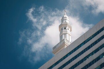 A white, slender tower with a spire, reminiscent of a steeple, rises above a modern building against a vibrant blue sky dotted with fluffy white clouds