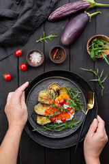 Hands holding a salad with fried eggplant, cream cheese, tomatoes and arugula on black wooden background top view, vertical photo. Vegetarian food