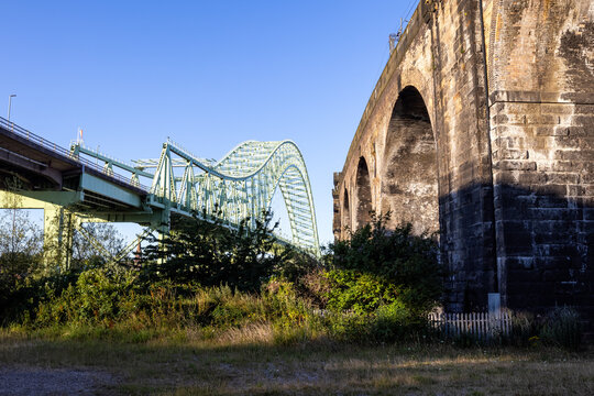 Stone railway viaduct alongside a steel arch road bridge, captured in warm early morning sunlight with shadows and clear blue sky - Powered by Adobe