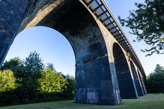 Low-angle view of a brick railway viaduct with multiple arches casting shadows, framed by trees and clear sky in early sunlight