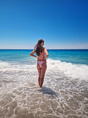 Rear view beautiful woman in bikini stands in the foamy waves of the clear blue sea, facing the wide horizon under a cloudless summer sky. Summertime vacation on a tropical beach, relaxation vibes