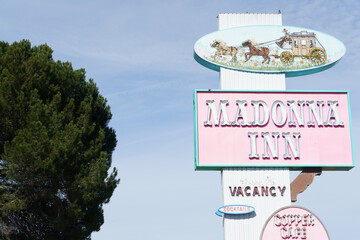 The iconic pink roadside sign for the Madonna Inn, featuring a vintage horse-drawn carriage illustration, stands tall against a bright blue sky.