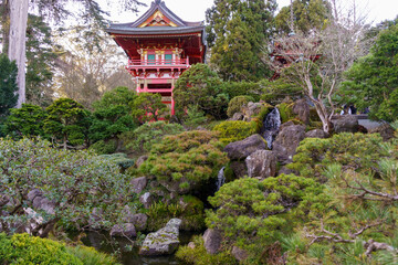 A vibrant red pagoda rises above a serene Japanese garden, where a waterfall tumbles over...