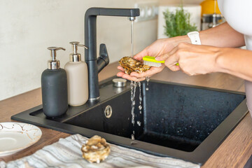 Female hands washing a fresh oyster under running tap water before opening. Hygiene and safety precautions when preparing shellfish at home.