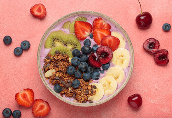 Vibrant and nutritious acai bowl topped with sliced kiwi, strawberries, blueberries, cherries, banana, and granola, a colorful and healthy meal or breakfast on a pink background