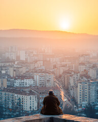Naklejka premium A person sits peacefully on a rooftop overlooking a city at sunset, embracing tranquility amid urban life.