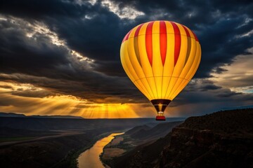 Obraz premium A vibrant yellow and red hot air balloon ascends over a dramatic landscape with a river at sunset, illuminated by golden sun rays