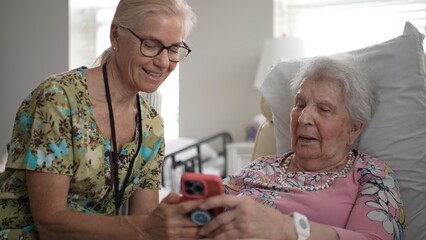 A compassionate nurse engages with an elderly woman by sharing photos and memories on a smartphone during a visit at a senior living facility.