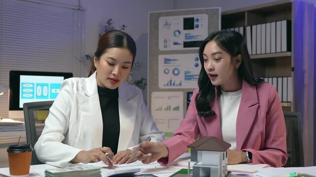 Professional real estate agent showing house model to her client, discussing mortgage and interest rates, pointing at documents and banknotes on the table