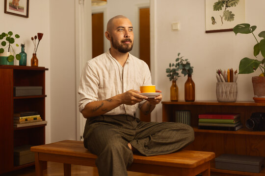Man sitting cross-legged on a bench, holding a tea cup in a cozy, stylish room. A calm moment of focus and relaxation.