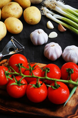 Fresh tomatoes, potatoes, green onions and garlic near vegetable peeler on dark background