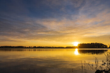 A peaceful sunset over a tranquil lake near Alkmaar, Netherlands, as the sky fades to soft hues of orange and pink, reflecting gently on the calm waters and surrounding countryside