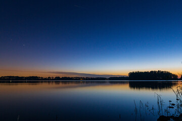 A peaceful sunset over a tranquil lake near Alkmaar, Netherlands, as the sky fades to soft hues of...