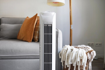 White tower fan against the background of a sofa and a basket with blankets in a modern light...
