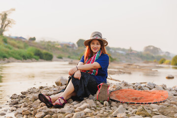 A smiling woman in a traditional hat rests on the pebbled riverbank with her fishing gear. A warm and inviting portrait showcasing the peaceful and happy side of a rural lifestyle.