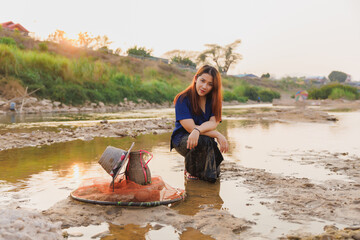 A woman in traditional clothing crouches in the river's shallows, her fishing gear beside her. A...