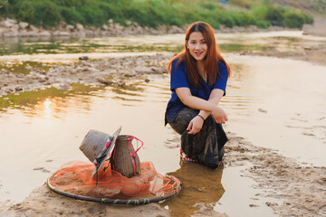 A smiling woman looks directly at the camera while crouching by her fishing gear on the riverbank....