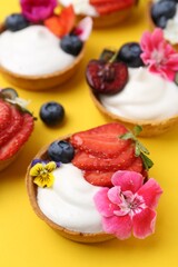 Sweet tartlets with berries and flowers on yellow background, closeup. Delicious dessert