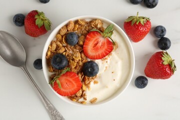 Tasty yogurt with granola, berries and spoon on white marble table, flat lay