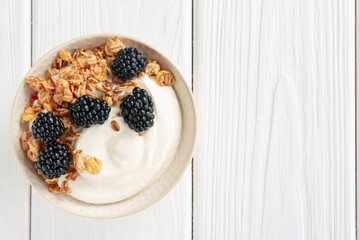 Tasty yogurt with granola and blackberries in bowl on white wooden table, top view. Space for text