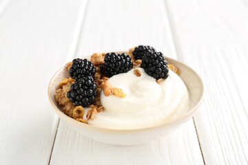 Tasty yogurt with granola and blackberries in bowl on white wooden table, closeup