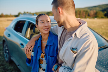 Young multi-ethnic couple enjoying their road trip, embracing joyfully near their car amidst the...